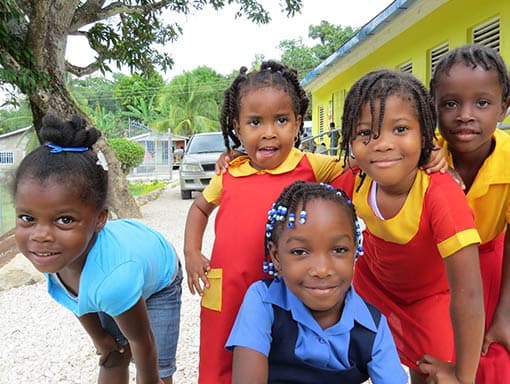 Five children in bright clothes gathered under a tree beside a yellow school building