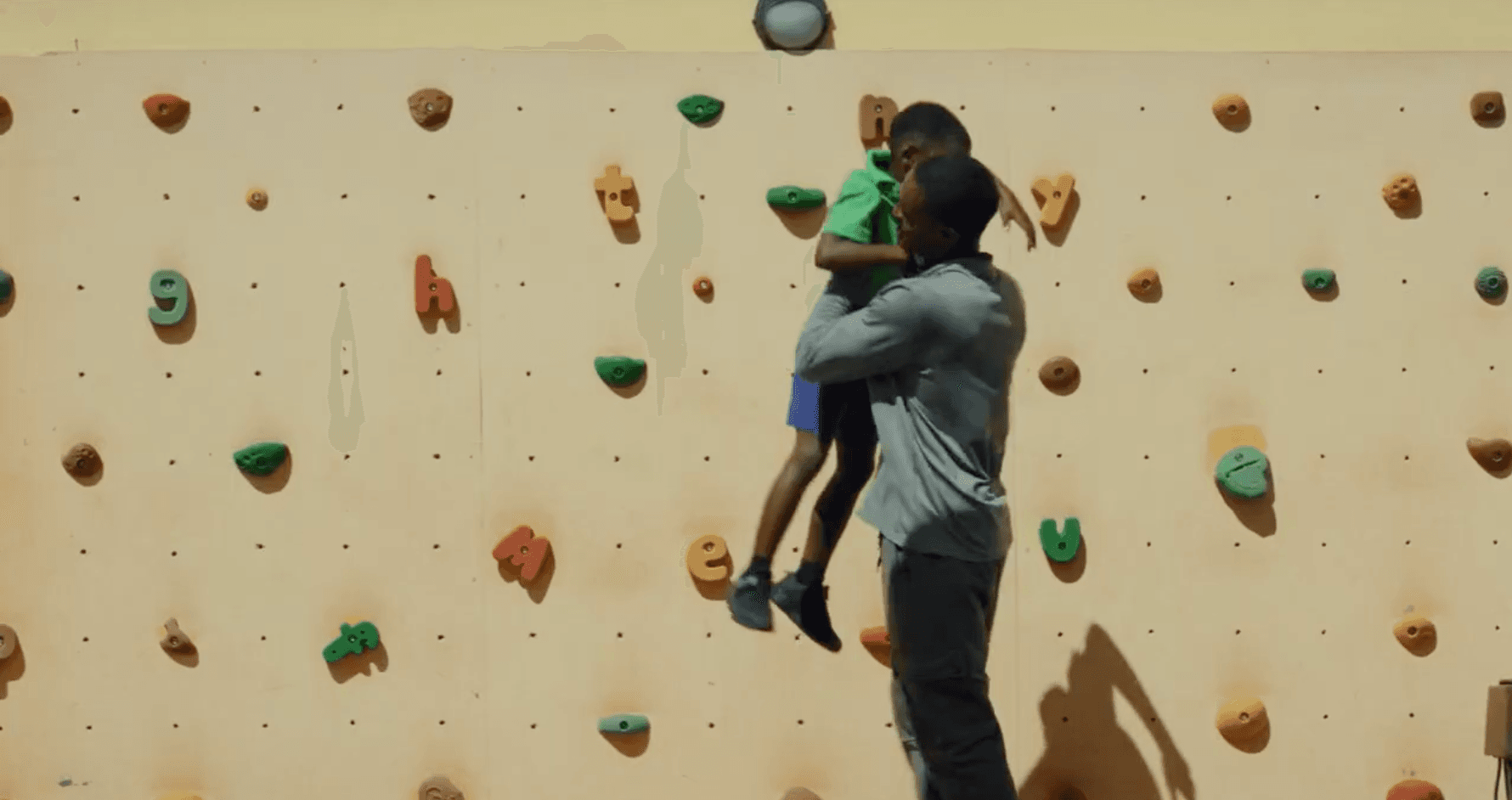 Image of a physical fitness coach helping a student climb a colorful rock wall at the Sav Inclusive School campus