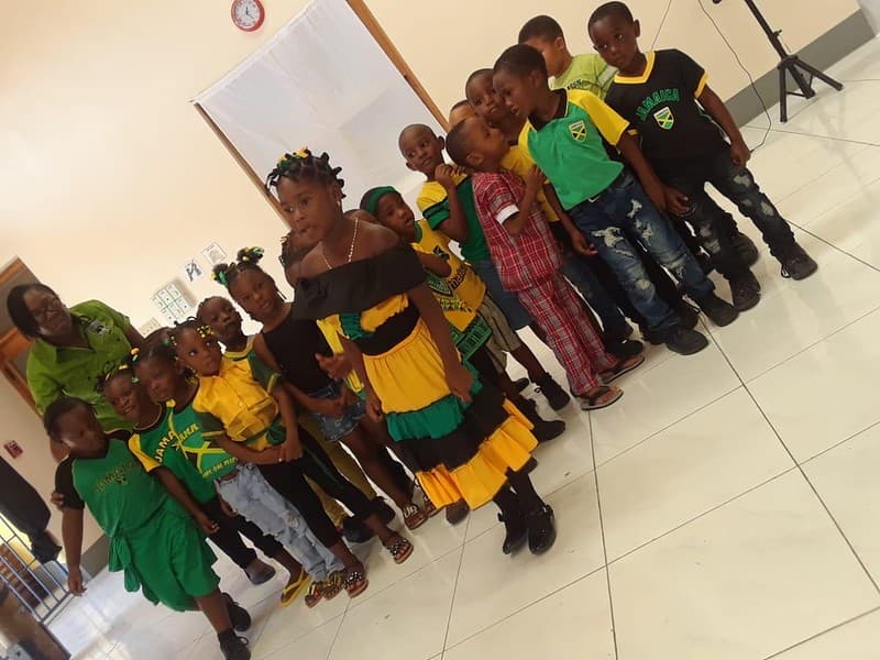 Around fifteen children in green and yellow uniforms lining up indoors, two teachers standing with them