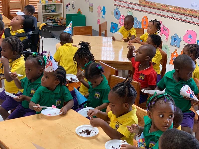 Children in party hats eating birthday cake at classroom tables, one of them seated in a support chair