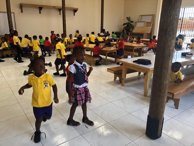 Young children in yellow and red uniforms between the long wooden tables of a tiled school hall