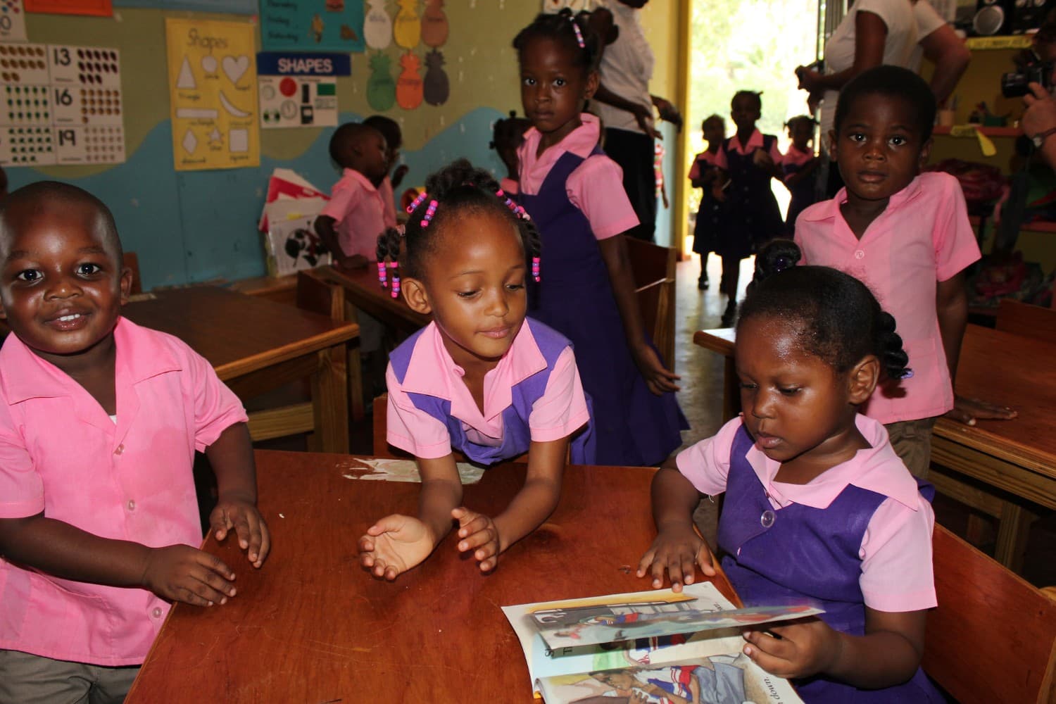Young children in pink and purple uniforms in a classroom hung with shapes and numbers charts