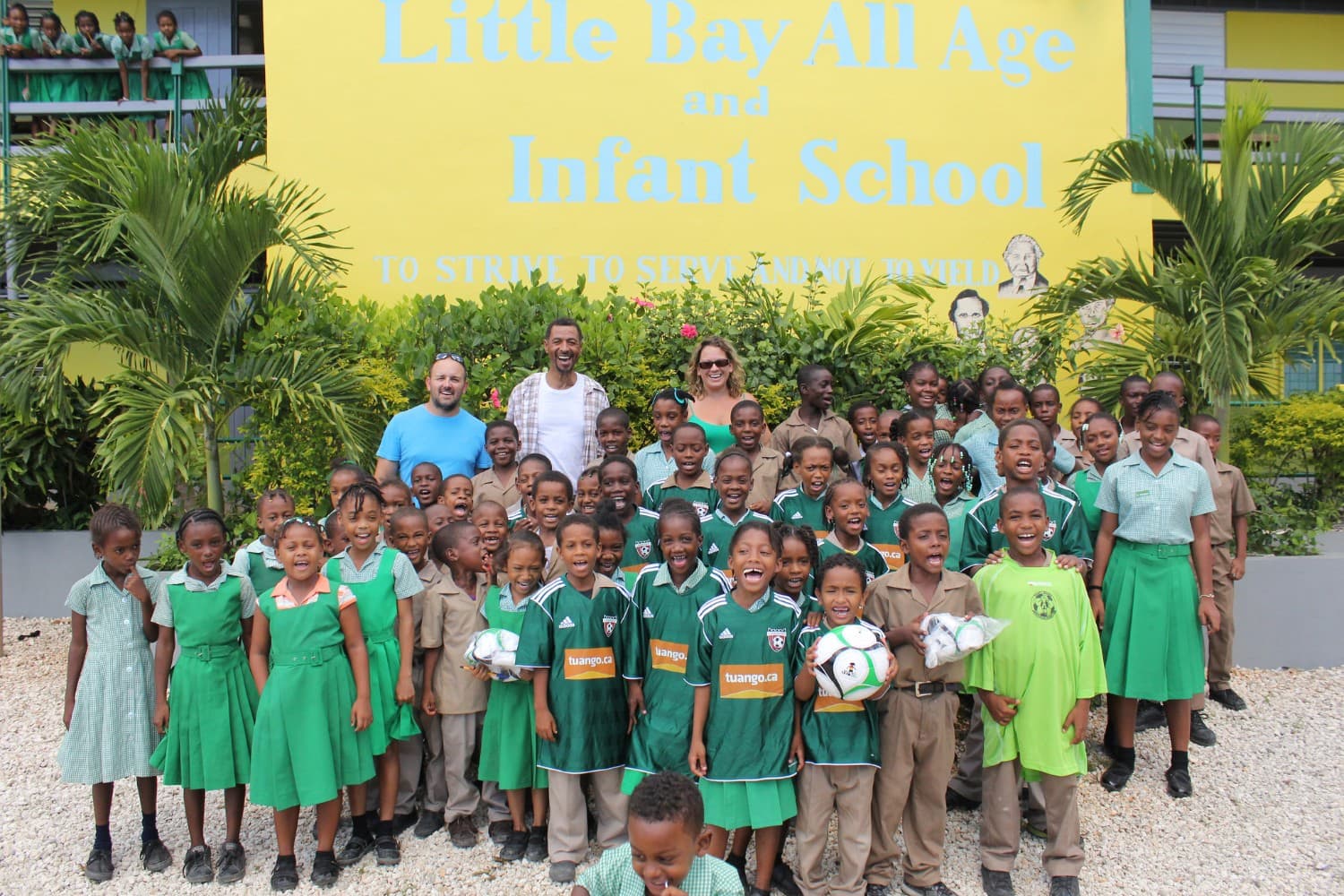 A whole school of children in green and khaki uniforms, several holding footballs, gathered with three adults under the Little Bay All Age and Infant School sign